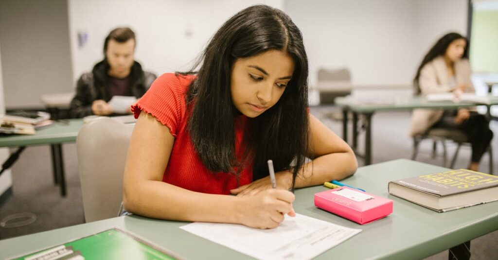 A college student concentrating while writing an exam in a classroom setting, surrounded by peers.