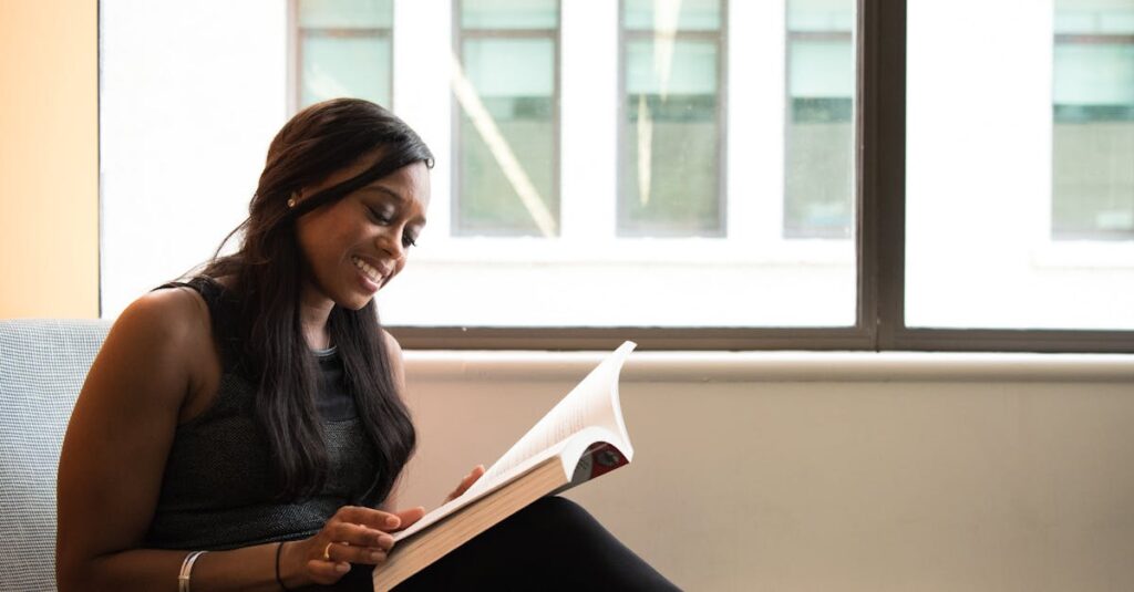 A smiling woman enjoys reading a book indoors by a large window, showcasing leisure and relaxation.