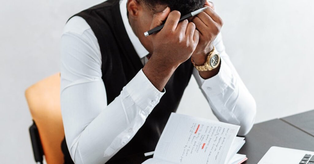 A businessman overwhelmed working indoors with a notebook, laptop, and hourglass.